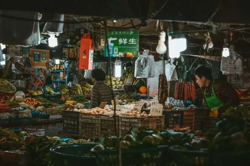 A brightly lit urban wet market stall at night, stacked with vegetables, eggs and cured sausage