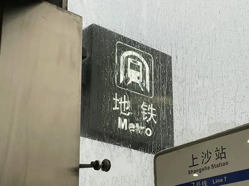 A weathered Shenzhen Metro sign reading '地铁 Metro' above a station nameplate for Shangsha Station, Line 7, rain streaking the glass