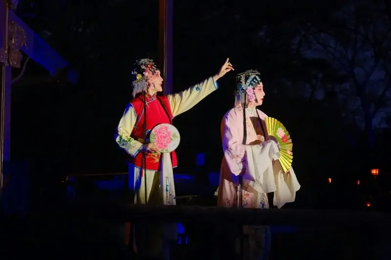 Two performers in elaborate Kunqu opera costume and headdress performing outdoors at night, lit by stage lighting