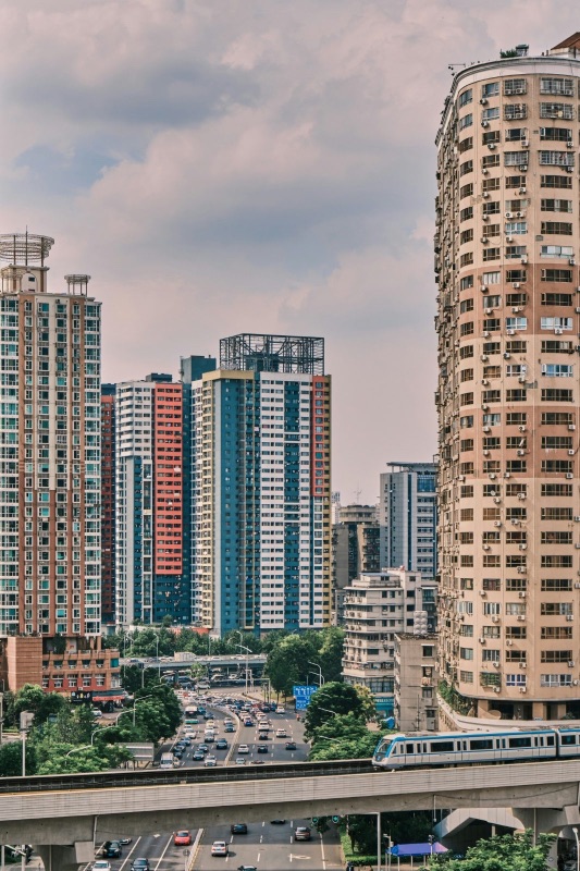 An elevated Wuhan Metro train running on a viaduct below a row of dense high-rise apartment towers, an overpass and traffic underneath