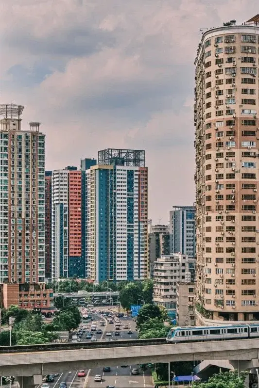 An elevated Wuhan Metro train running on a viaduct below a row of dense high-rise apartment towers, an overpass and traffic underneath