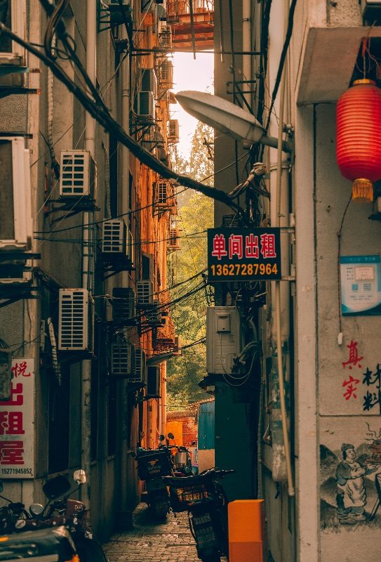 A narrow Wuhan alley lined with air-conditioning units and a red sign advertising a single room for rent, a red lantern hanging nearby