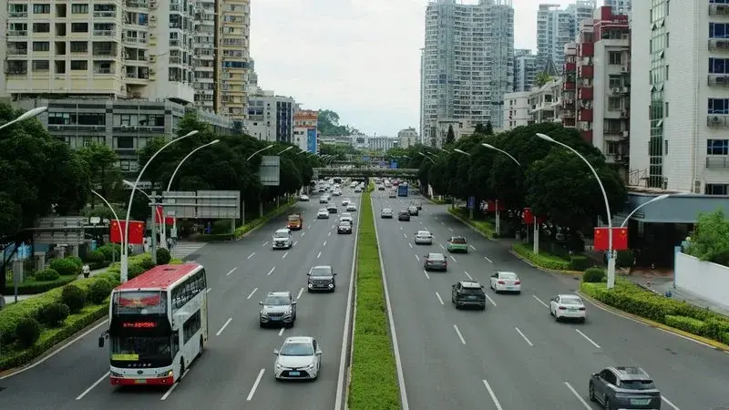 A wide tree-lined boulevard in Xiamen with a double-decker bus and traffic under an overcast sky, apartment towers on both sides