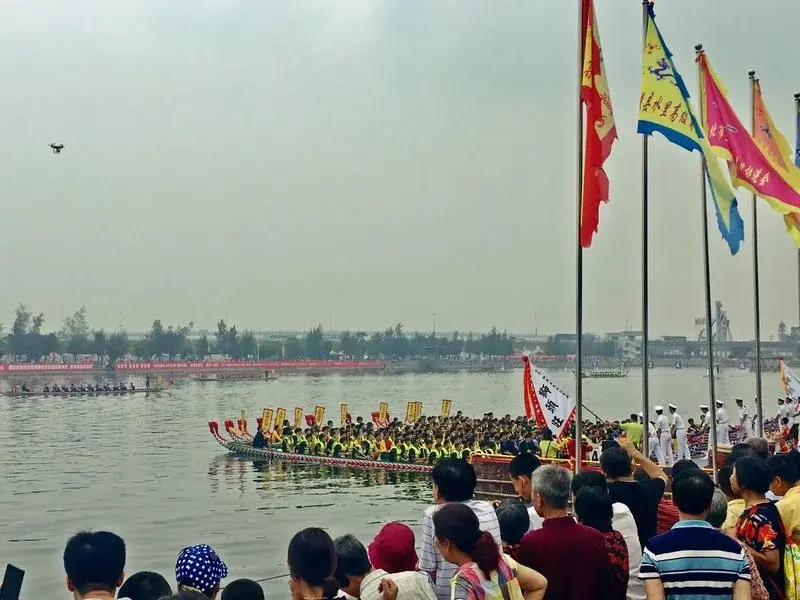 A dragon boat race in Xiamen, with colorful flags and a crowd of spectators watching from the shore