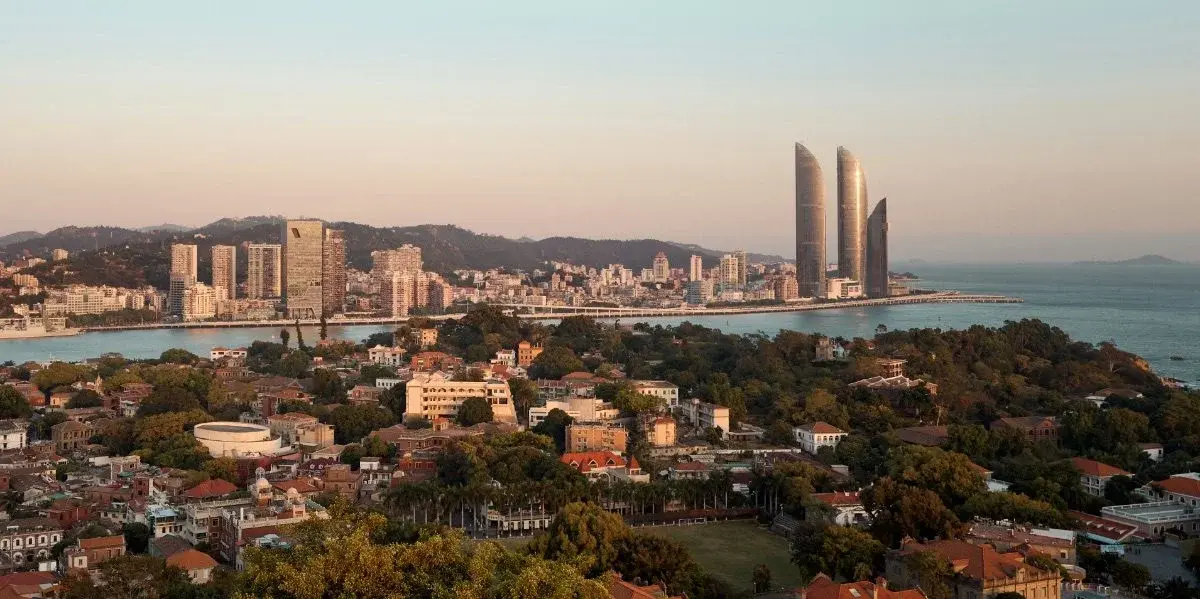 Xiamen skyline at dusk, Gulangyu's colonial-era red-roofed buildings in the foreground and the twin Xiamen International Centre towers rising across the water