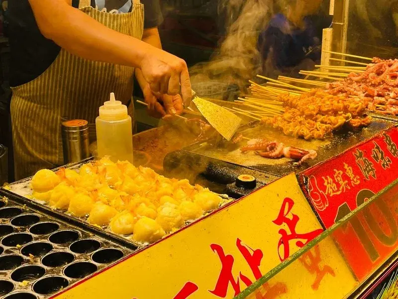 A street food vendor grilling seafood skewers and octopus at a busy night market stall