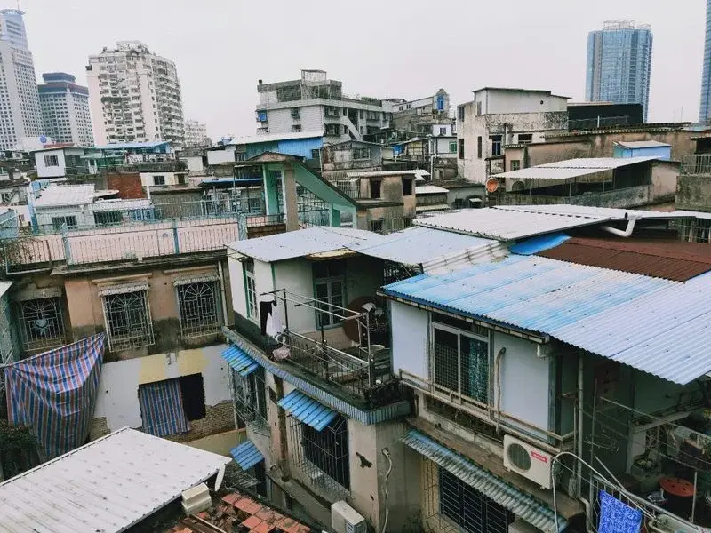 A dense rooftop view of older low-rise apartment buildings with makeshift extensions and air conditioning units, high-rises visible in the distance