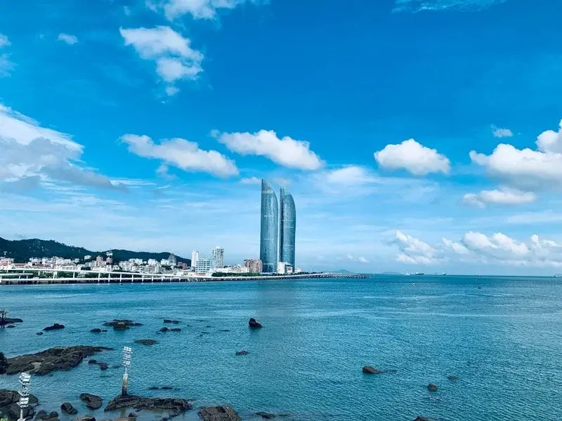 Xiamen's twin International Centre towers rising from the sea under a clear blue sky, seen from a rocky waterfront