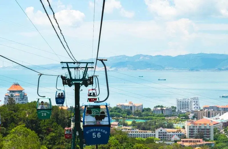 Cable car gondolas ascending over a hillside with the sea and distant coastline visible below