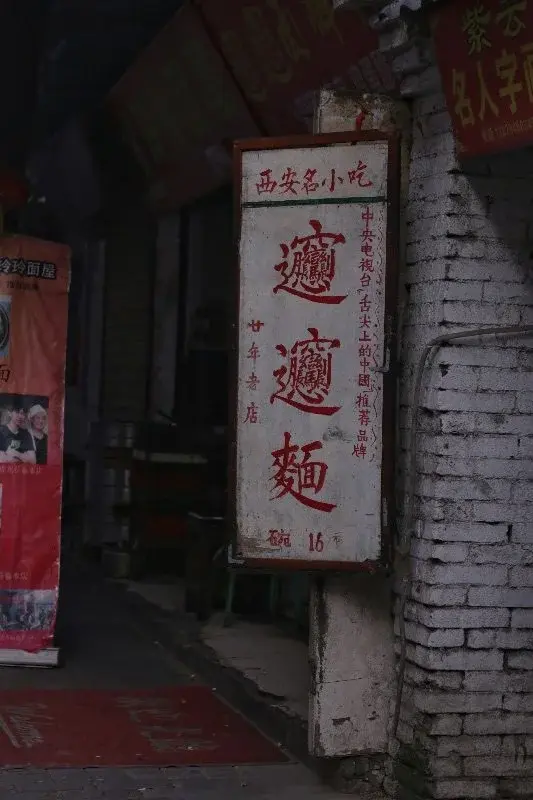 A weathered hand-painted restaurant sign in Xi'an reading '西安名小吃' with the invented biángbiáng character below it, priced at ¥16 a bowl