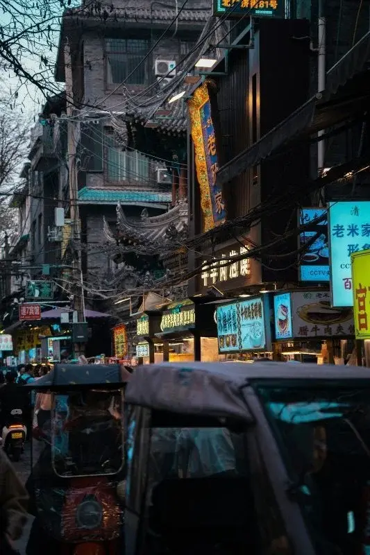A dimly lit Muslim Quarter street at dusk, restaurant signs and lanterns crowding overhead, a covered cart in the foreground