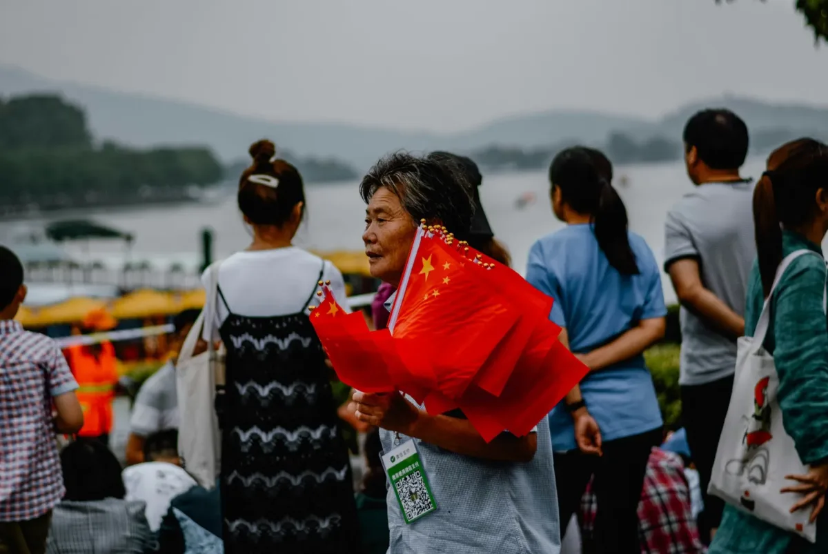 A flag seller carrying a bundle of small Chinese flags along a lakeside promenade on an overcast morning, crowds strolling behind her