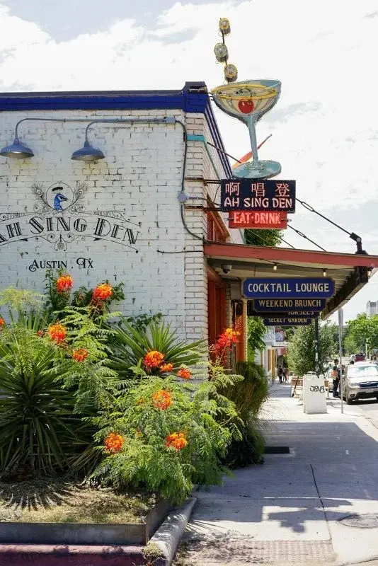 An Austin cocktail lounge storefront with a Chinese-character sign reading 'Ah Sing Den' beside blooming flowers