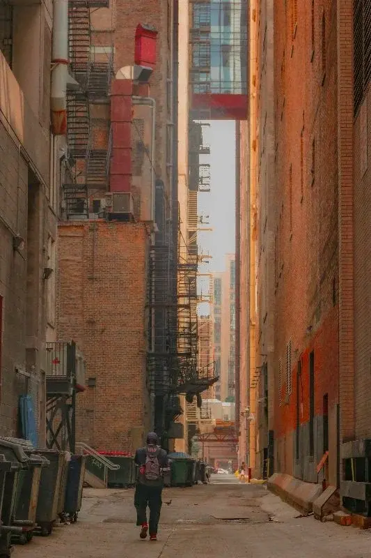 A narrow Chicago service alley between brick buildings, fire escapes running up the walls, a pedestrian walking past the trash bins
