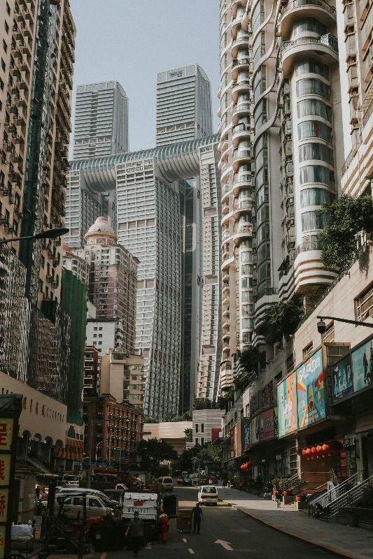 A canyon of Chongqing high-rise towers seen from street level, including a curved connecting structure joining two towers, with red lanterns visible on a shopfront below