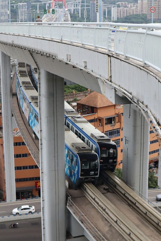 A Chongqing monorail train on an elevated curved viaduct threading between buildings, with a road bridge running above it