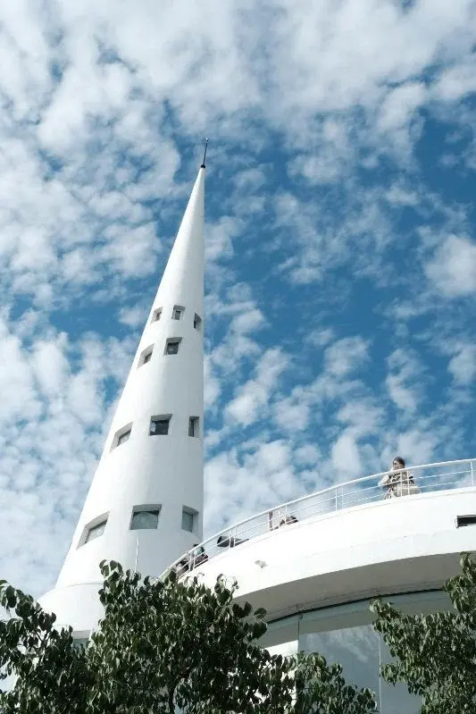 A tall, tapering white tower with small square windows against a bright blue sky, part of a building's observation platform