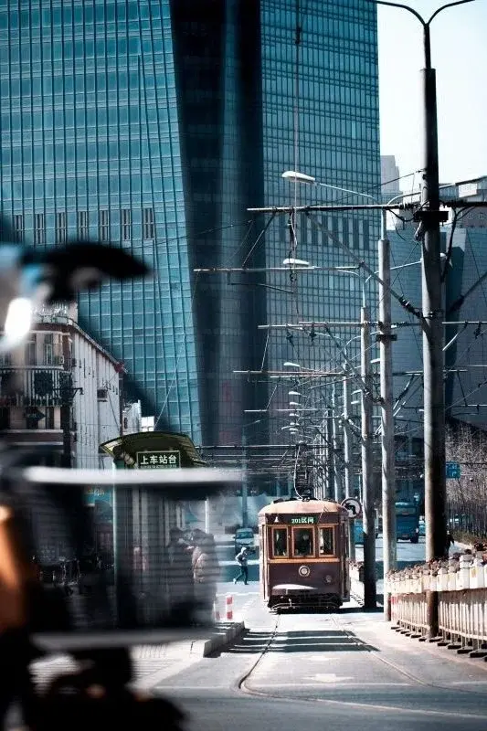 A Dalian tram car running on a street toward a glass office tower, with a green boarding-platform sign visible