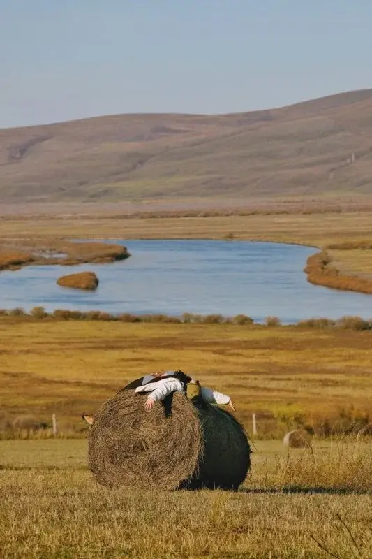 A person lying atop a large hay bale beside a winding river on the grassland near Hohhot