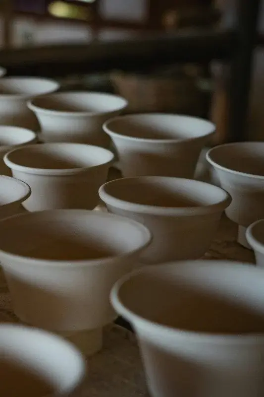 Rows of unfired, pale clay cups drying on a table in a Jingdezhen workshop, awaiting their turn in the kiln