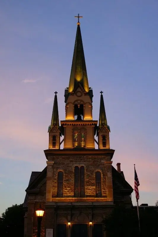 A church steeple in Minneapolis lit gold at dusk against a purple sky, with an American flag beside it