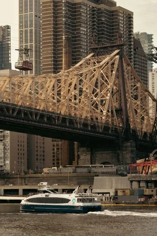 The Roosevelt Island Tramway crossing beneath the Queensboro Bridge, with a ferry passing on the East River below