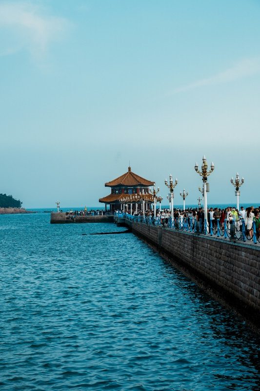 Visitors walking along Qingdao's Zhan Qiao pier toward its pavilion, lampposts lining the railing