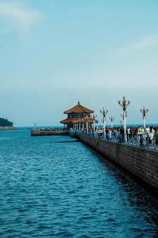 Visitors walking along Qingdao's Zhan Qiao pier toward its pavilion, lampposts lining the railing