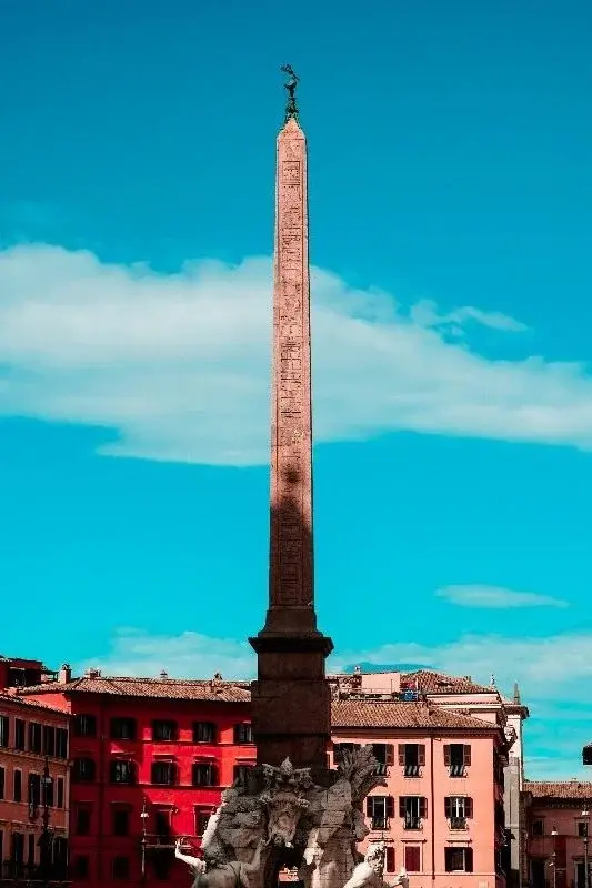 An ancient Egyptian obelisk rising above a Roman piazza, red and pink buildings in the background