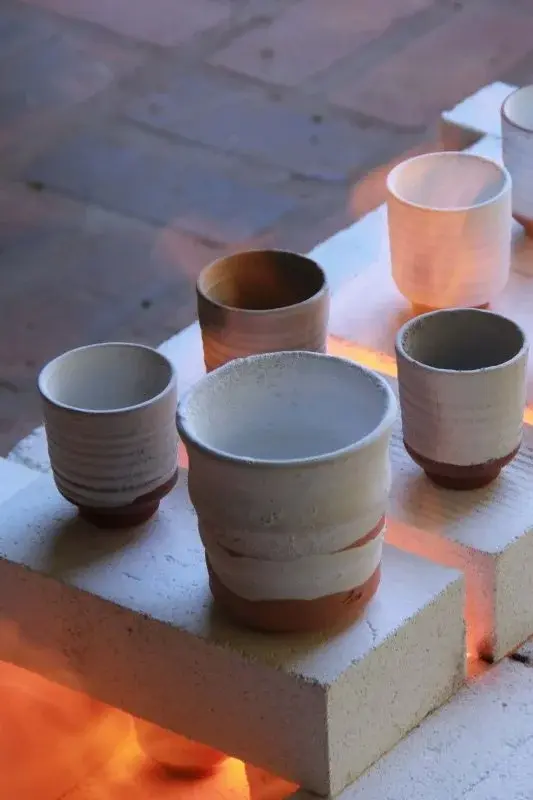 Freshly fired white-and-terracotta cups arranged on kiln bricks, the glow of the kiln fire showing through the gaps