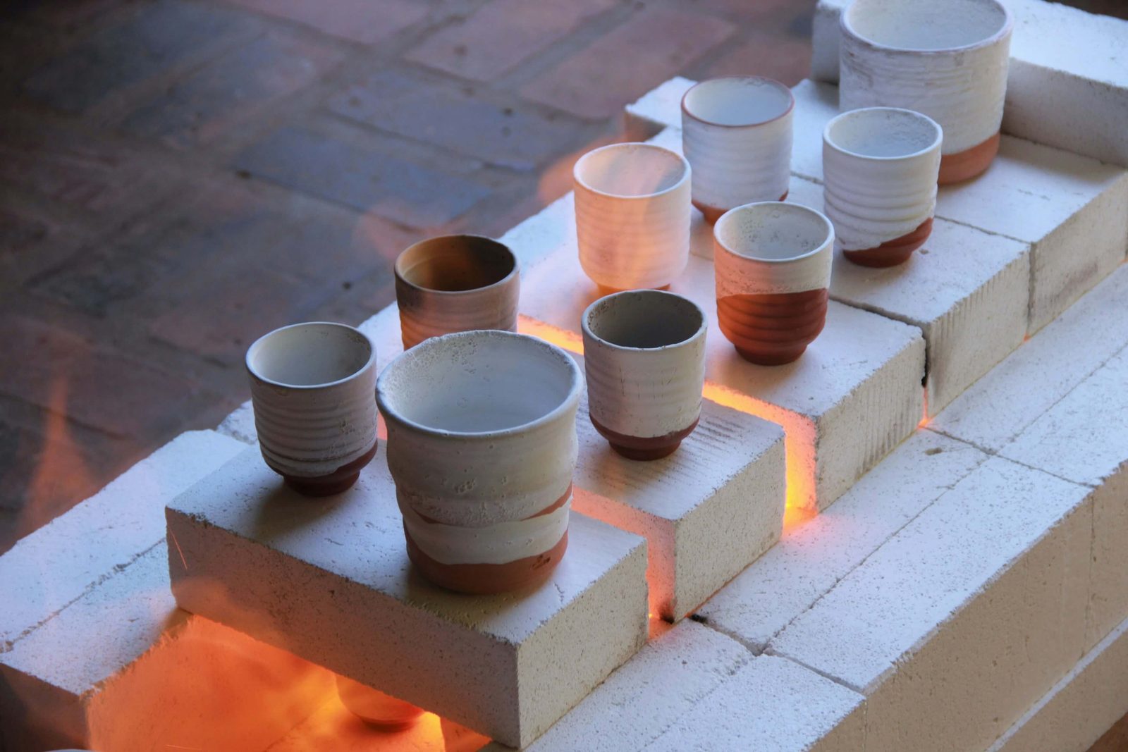 Freshly fired white-and-terracotta cups arranged on kiln bricks, the glow of the kiln fire showing through the gaps
