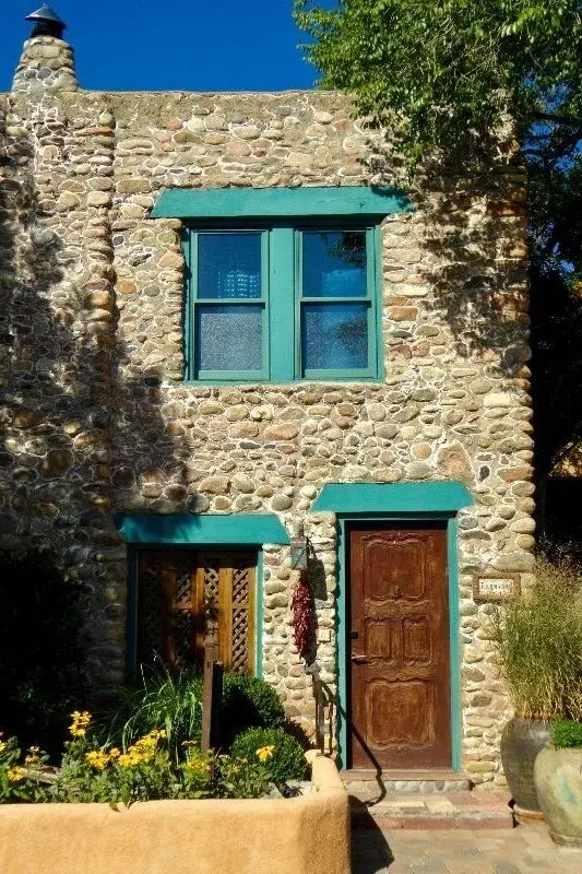 A river-stone Santa Fe house with turquoise-trimmed windows and door, potted plants and flowers out front