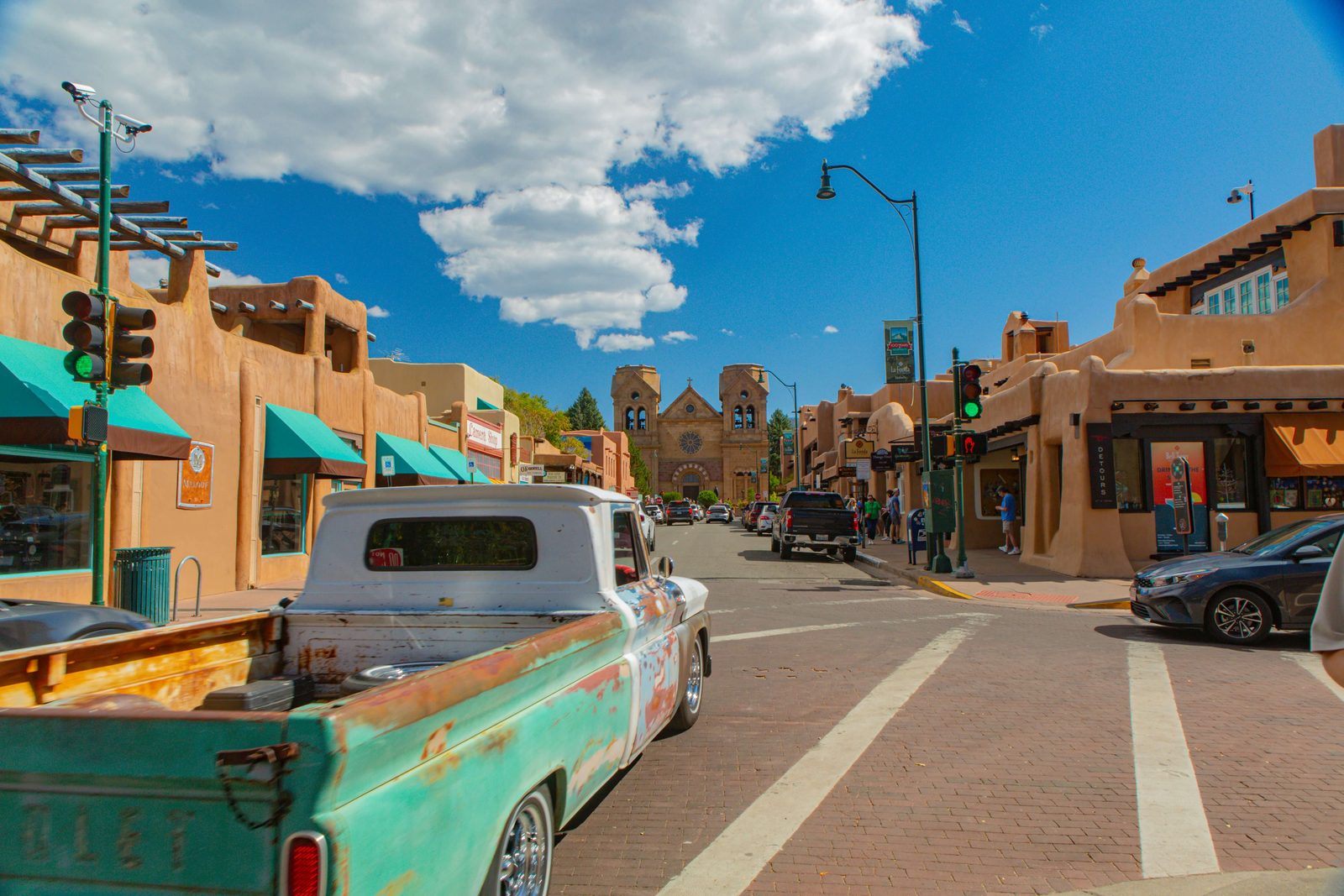 A Santa Fe street of adobe buildings with turquoise awnings, a vintage pickup truck, and the stone cathedral at the end under a blue sky