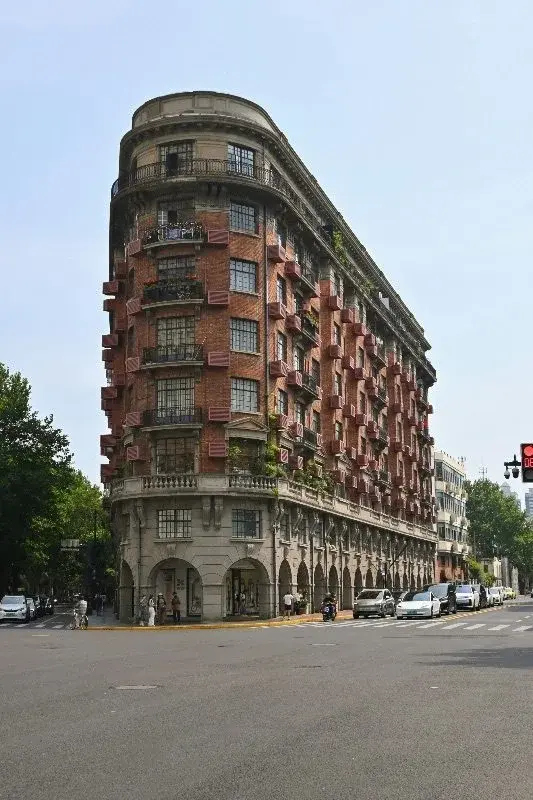 A rounded-corner early 20th-century brick apartment building on a Shanghai street corner, with balconies and shopfronts at street level