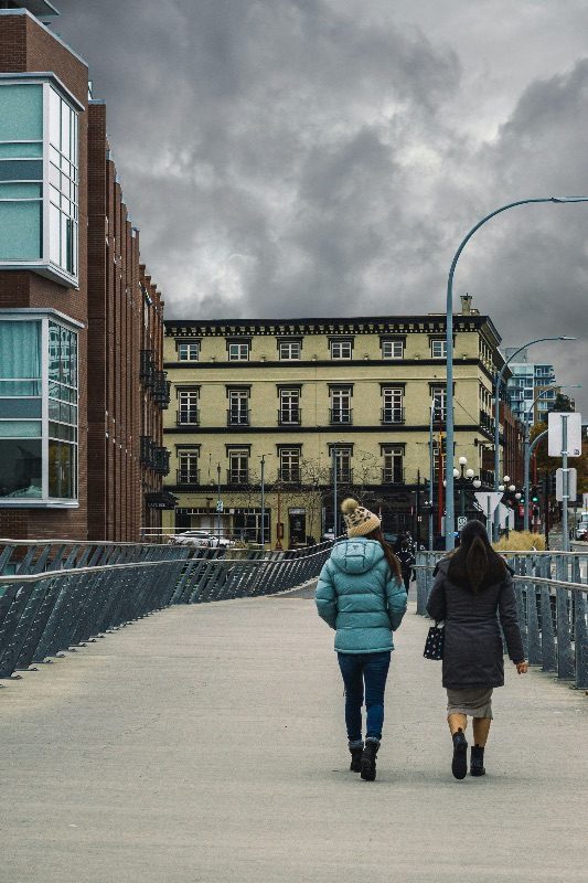 Two people walking away from the camera on a pedestrian bridge in Victoria under an overcast sky, old brick buildings ahead