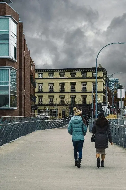 Two people walking away from the camera on a pedestrian bridge in Victoria under an overcast sky, old brick buildings ahead