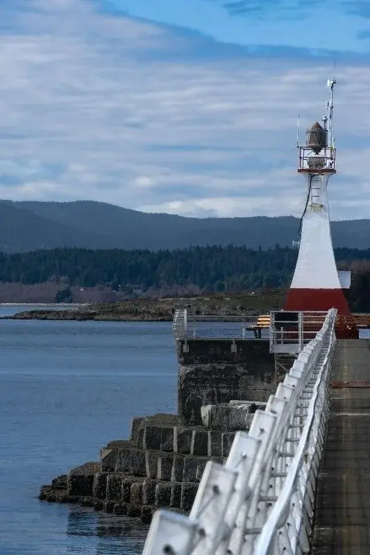 A small red-and-white lighthouse at the end of Victoria's Ogden Point breakwater, forested hills across the water