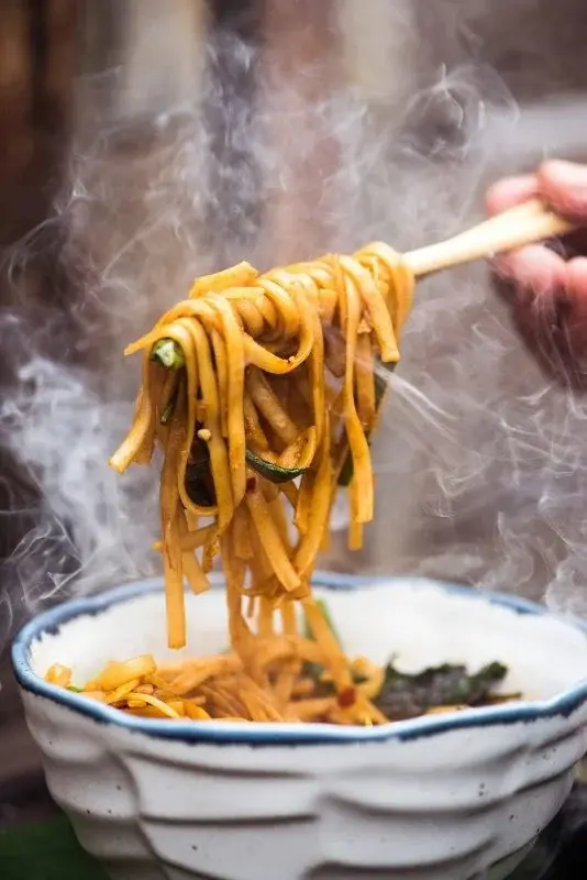Chopsticks lifting a steaming tangle of chili-red hand-pulled noodles from a bowl