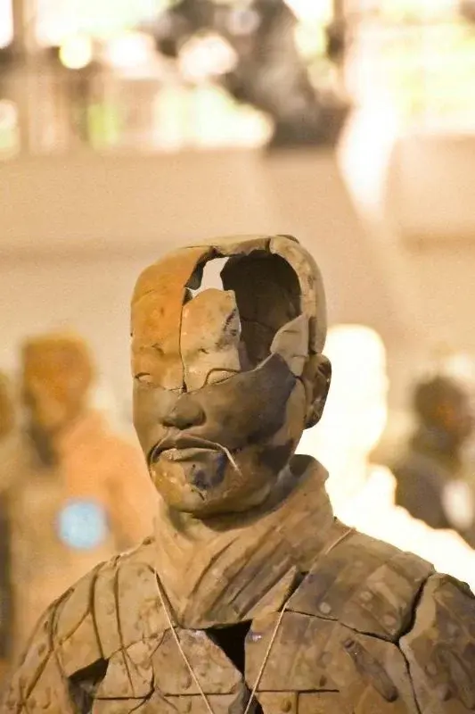 A cracked, weathered terracotta warrior head on display, other soldiers visible blurred in the background