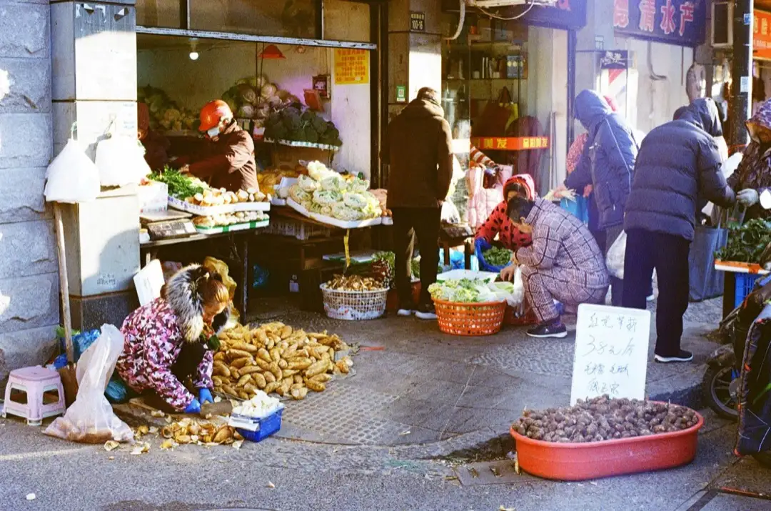 Street vegetable stall on a winter morning in a Chinese neighbourhood, shoppers in thick coats buying greens and root vegetables laid out on the sidewalk under a shop awning