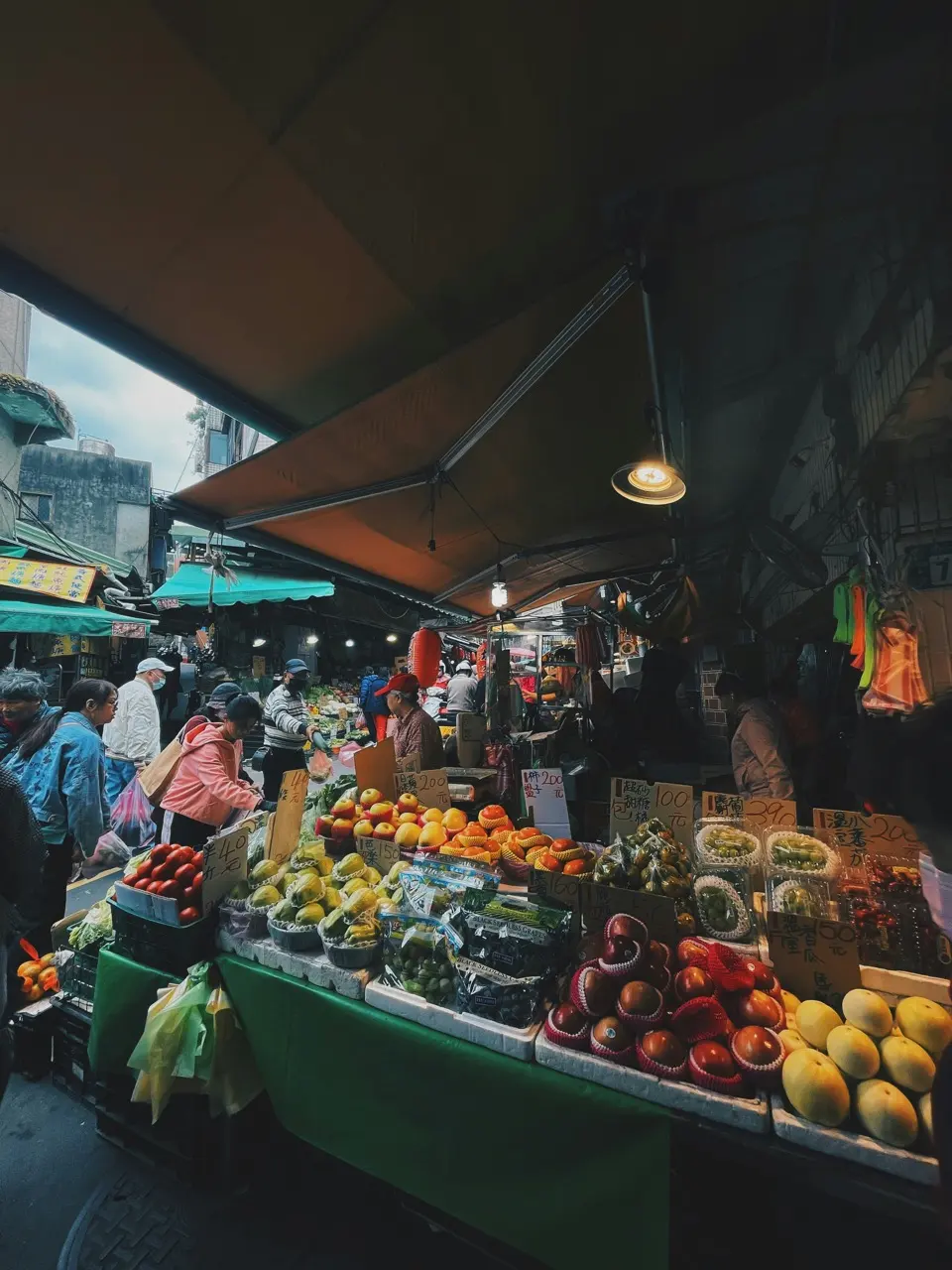 Covered street market in southern China with orange awnings, fruit stalls piled with grapes and mangoes, and shoppers browsing under hanging lamps