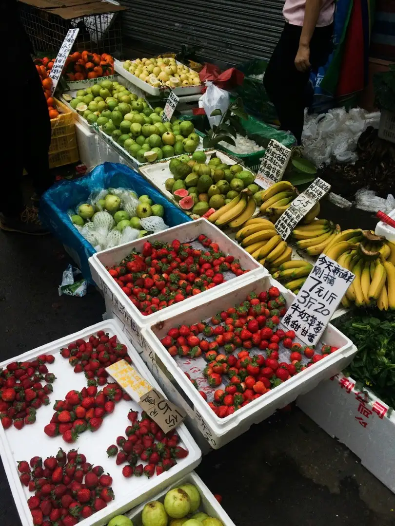 Morning market stall with boxes of fresh strawberries, bananas and guavas, and handwritten Chinese price signs quoting yuan per jin