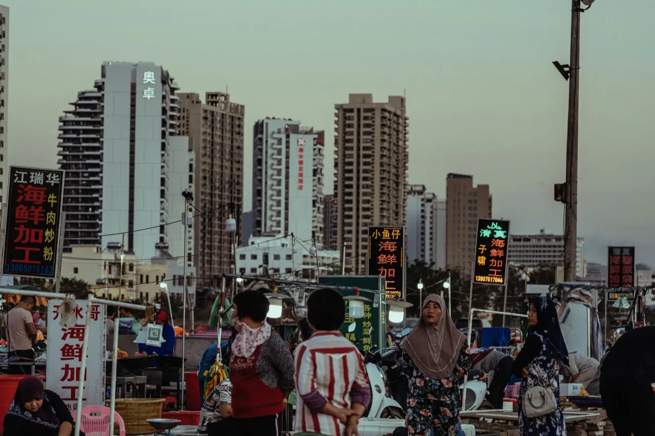 Outdoor seafood market at dusk in Sanya, lit signs advertising seafood processing and rice noodle soup in front of residential high-rise towers