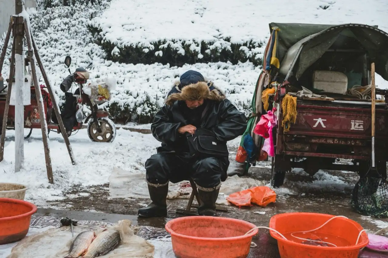 Fish vendor in a heavy winter coat sitting at an open-air roadside stall on a snowy day, live fish and shellfish in orange plastic basins, a snow-covered slope and a motorised cargo tricycle behind him