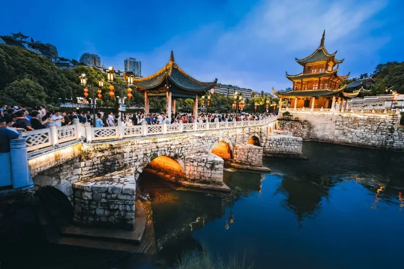 Crowds walking across the historic stone bridge at Jiaxiu Pavilion in Guiyang during a cool summer evening, with traditional architecture lit up against the blue twilight sky