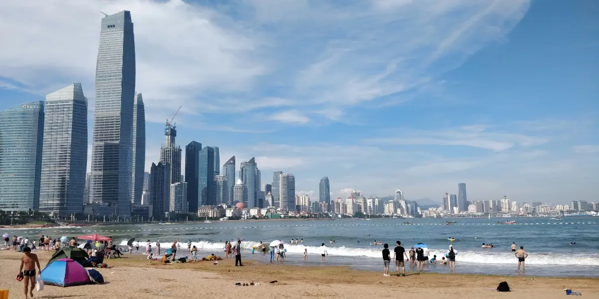 Crowded beach in Qingdao with skyscrapers in the background, people swimming and sunbathing on a hot summer day.