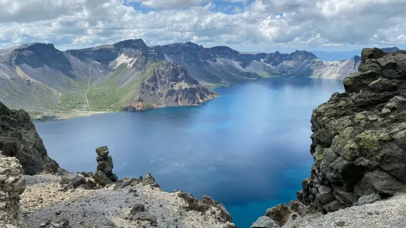 Panoramic view of the Heaven Lake at Changbai Mountain in summer, featuring deep blue water surrounded by volcanic peaks and rocky terrain under a cloudy sky