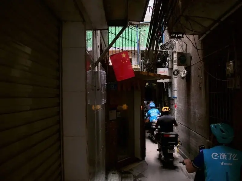A narrow residential alley with a small Chinese flag hanging overhead, two delivery riders on scooters heading down it