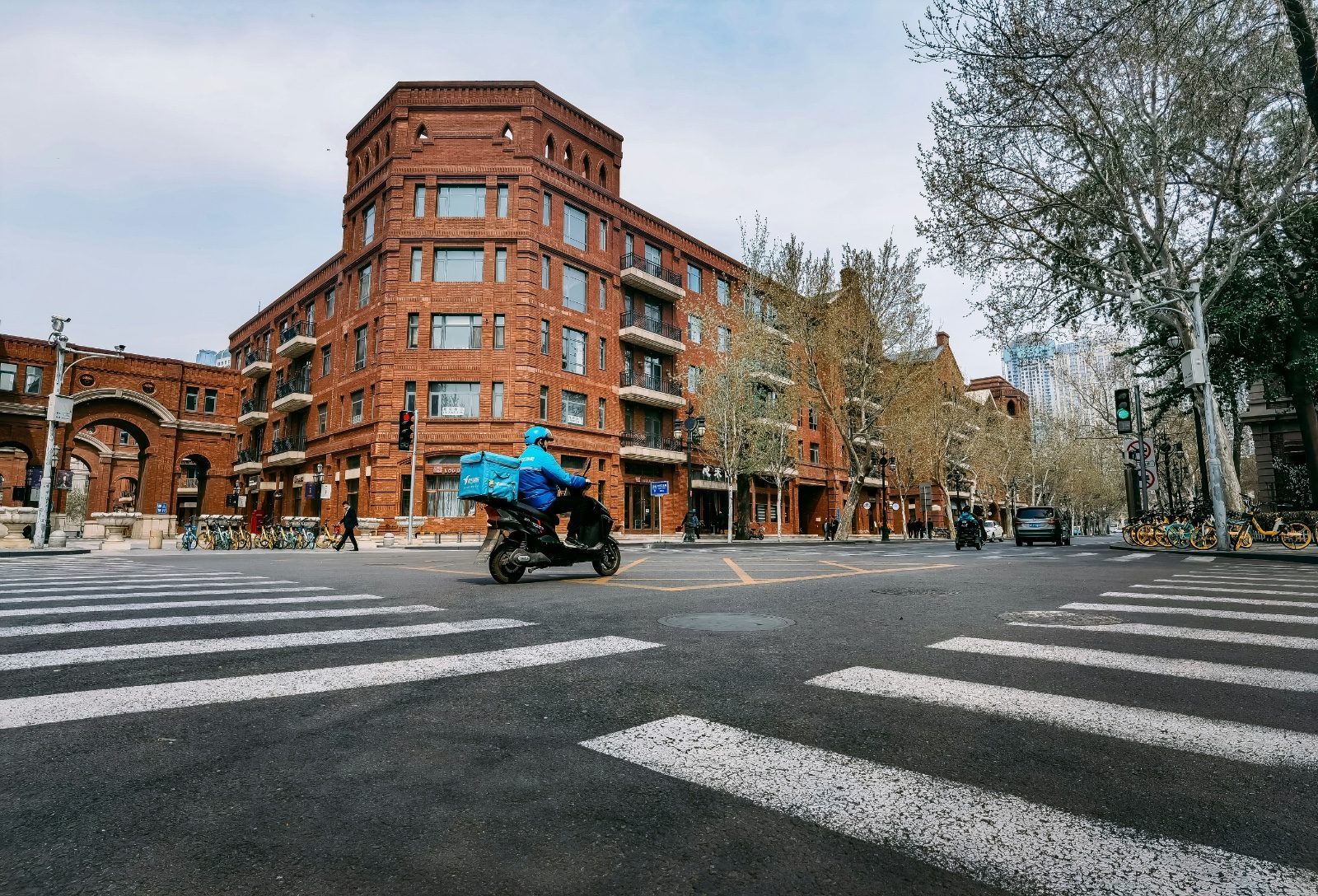 An Ele.me rider in a blue uniform riding through a street lined with red-brick colonial-era buildings