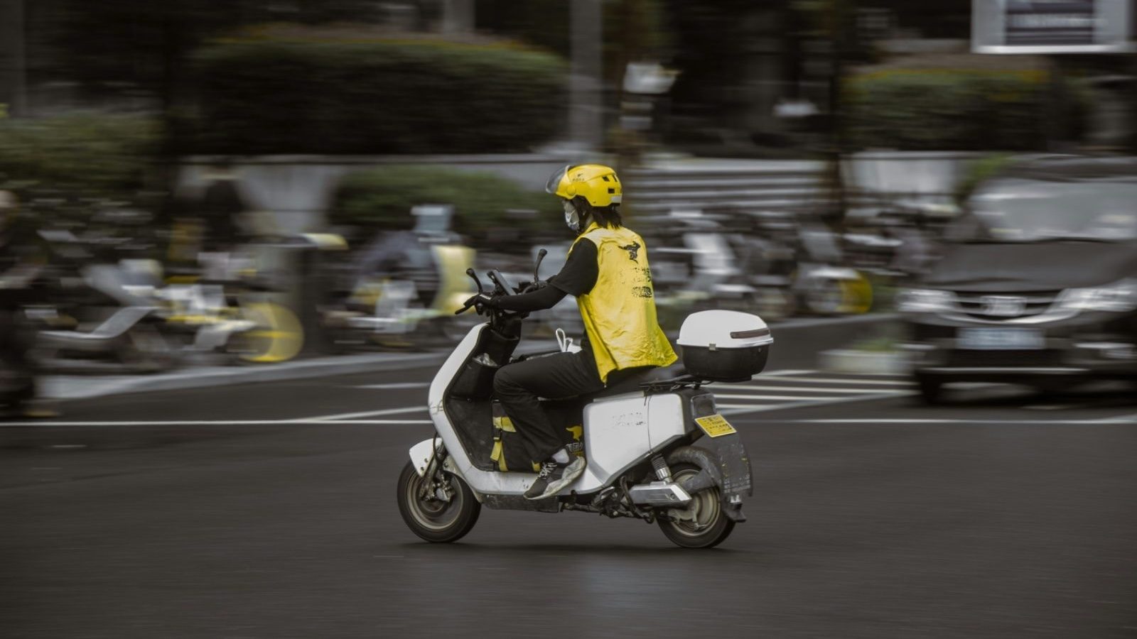 A delivery rider on a scooter, motion-blurred, weaving through daytime city traffic